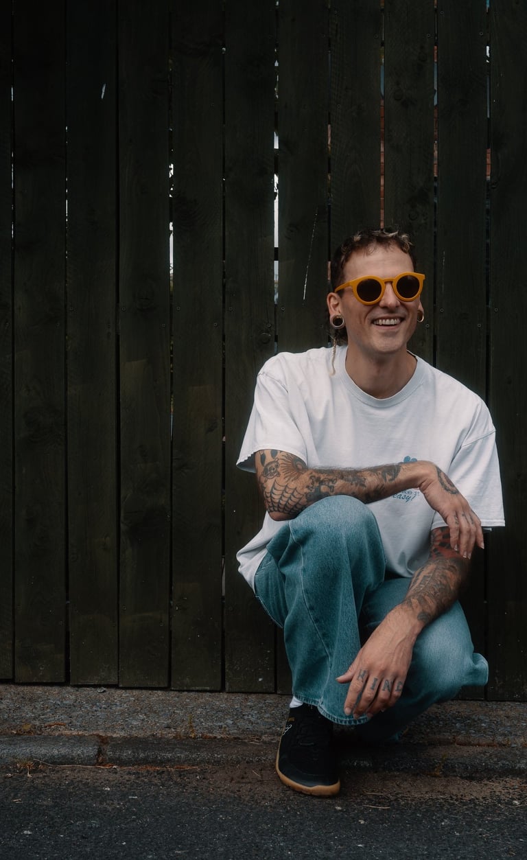 Cody with yellow sunglasses, a baggy t shirt, and baggy jeans, crouched in front of a green fence.