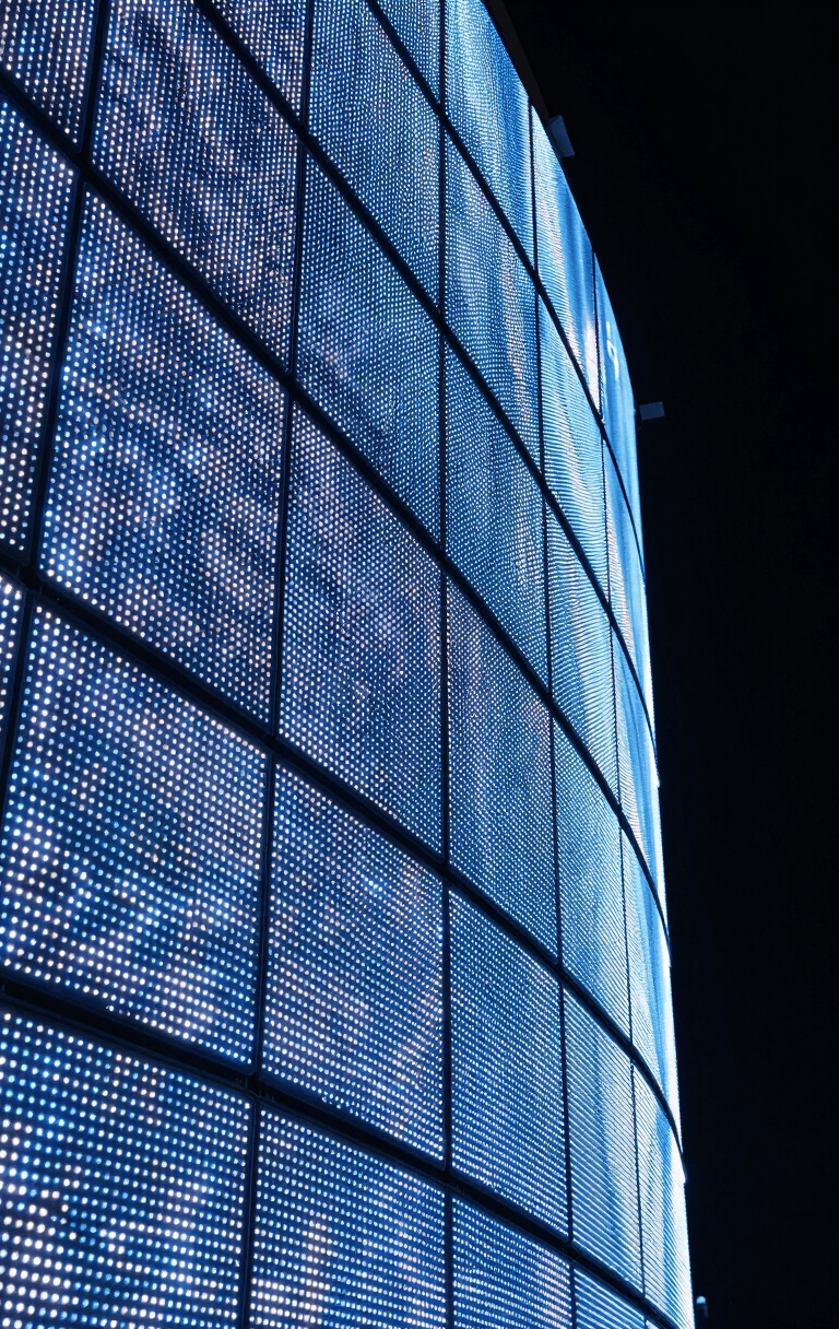 Extreme close-up of a large curved LED panel array in a darkened installation space, electric blue and white LED pixels illuminating the precise seam geometry and curve radius along the panel's edge, nighttime controlled lighting, no ambient fill, architectural framing from a low oblique angle showing depth and curvature
