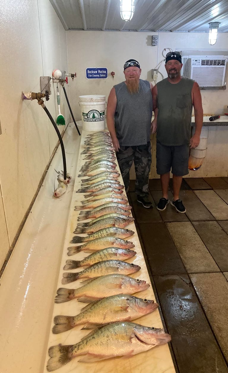 Two smiling fishermen standing by a long row of fresh-caught crappie at a marina fish cleaning station.