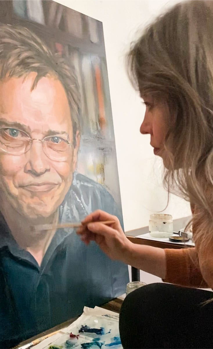 Stefanie placing final touches on oil portrait painting of smiling man with glasses.