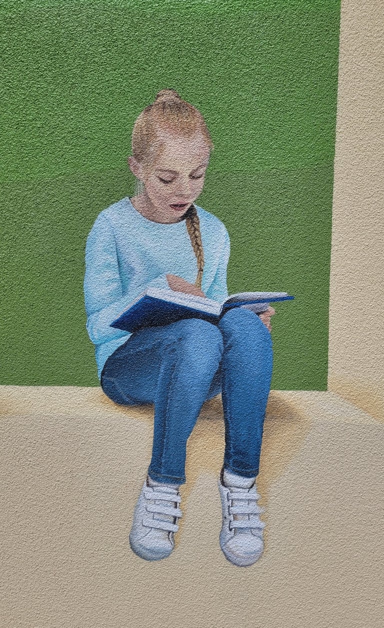 Fresque peinte sur façade. Une fille lit assise sur le bord d'un muret.