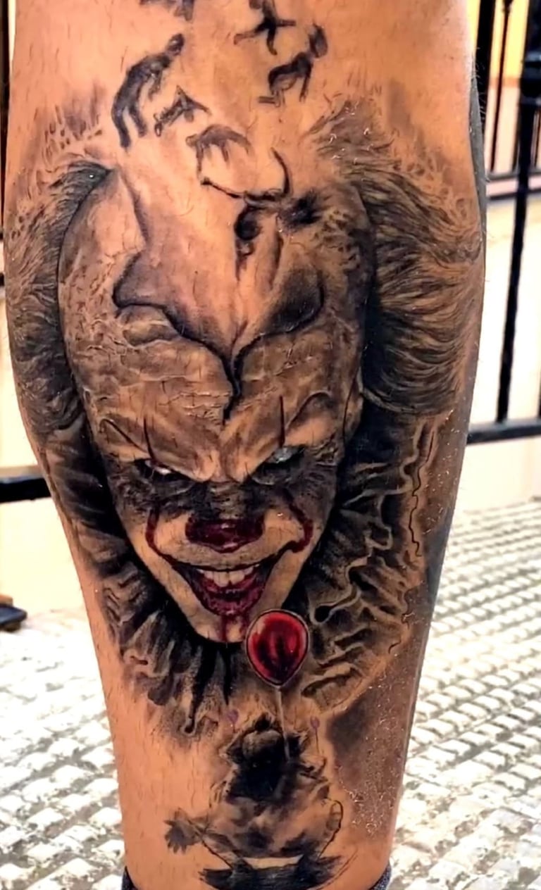 A realistic black and grey Pennywise the clown tattoo with a red balloon on a person's leg.