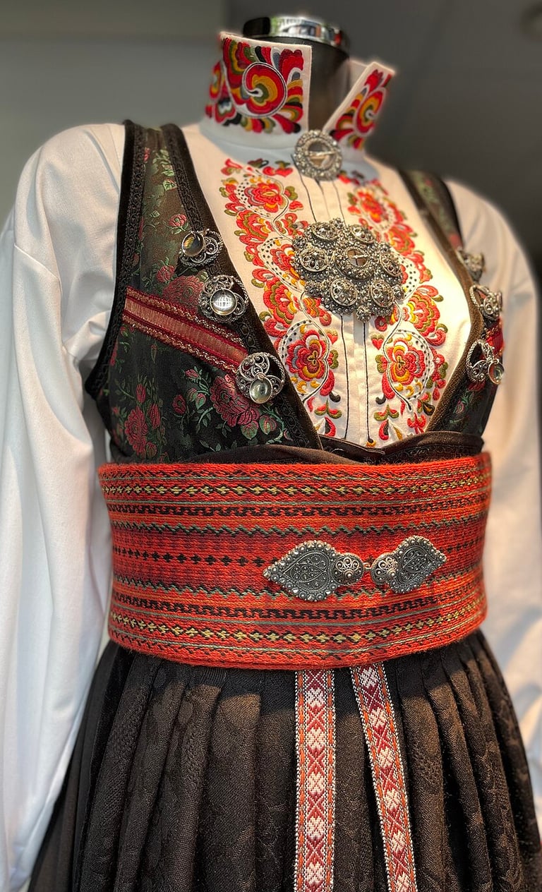 Black Bunad with a wide red belt and embroidered white shirt on a mannequin