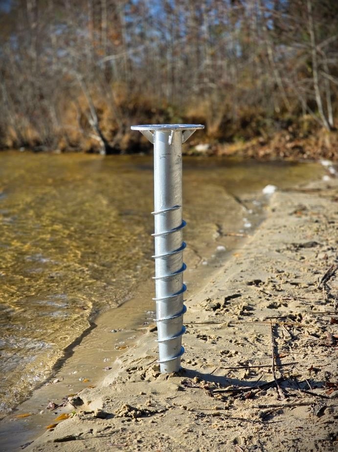 A galvanized steel screw pile foundation installed in the sandy shore of a lake for dock construction.
