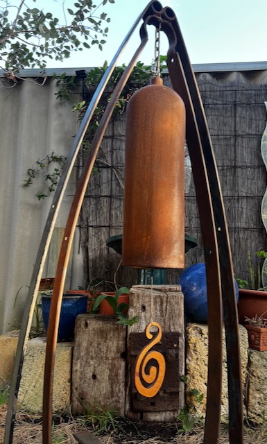 Rustic metal garden bell hanging from a tripod stand in a backyard garden setting.