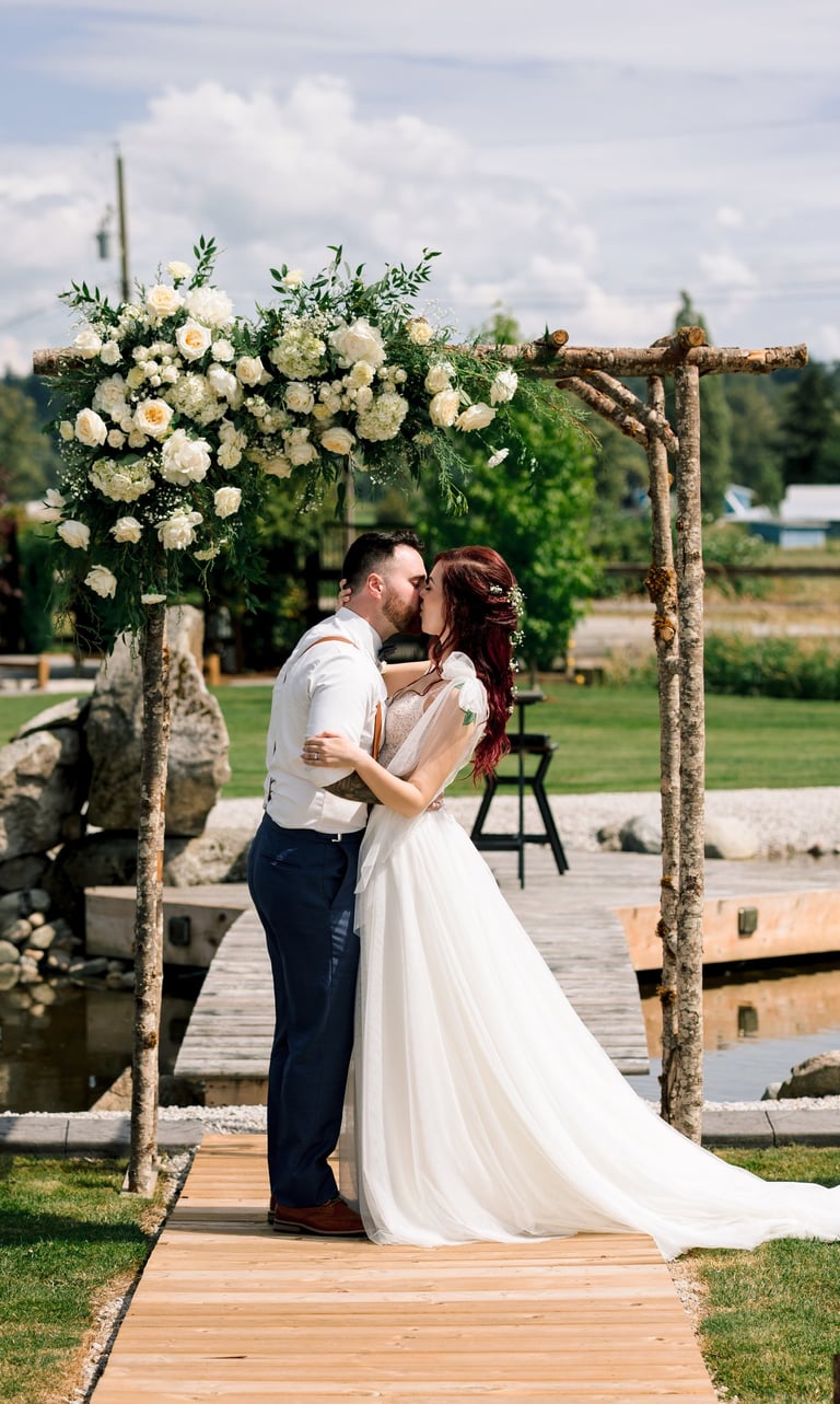 Newlywed couple kissing under a rustic wooden wedding arch decorated with white roses at an outdoor ceremony.