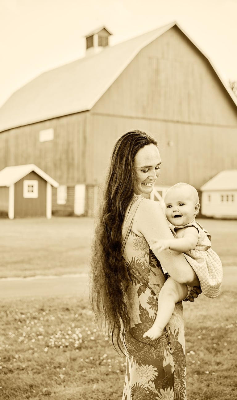 Sepia photograph of a smiling mother holding her happy baby in front of a large rural farm barn.