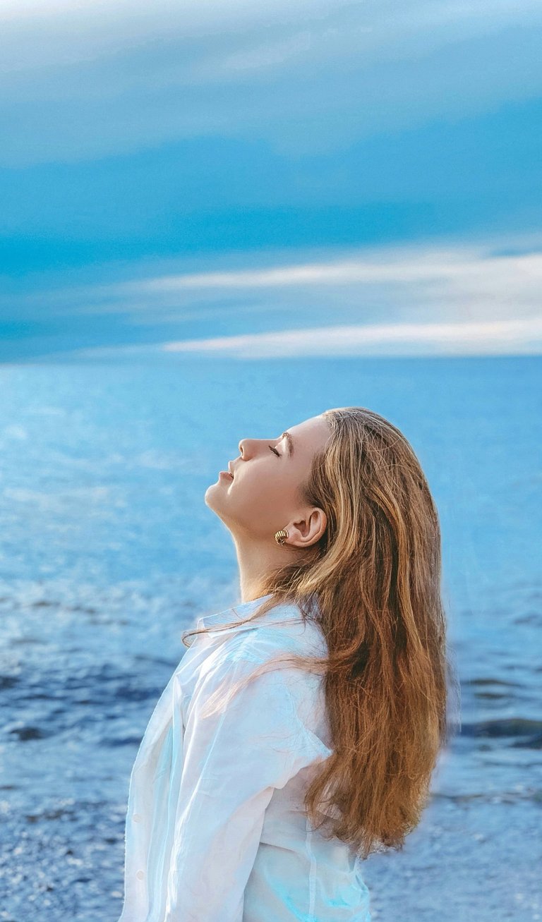 A young woman with long hair enjoys a peaceful moment by the blue ocean waves under a cloudy sky.