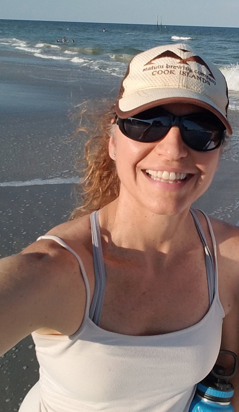 A smiling woman wearing sunglasses and a baseball hat takes a selfie on a sunny beach.
