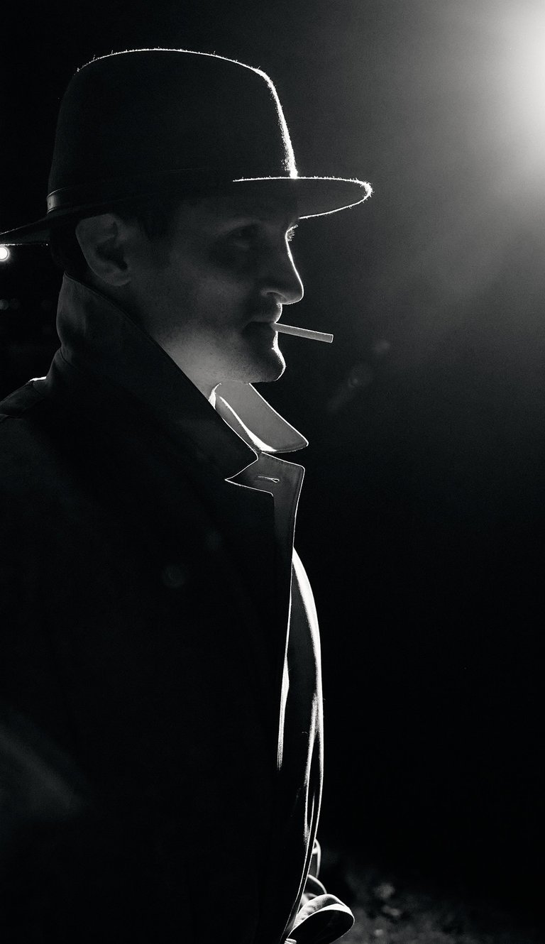 a man in a hat and a cigarette standing a dark area with street light from afar
