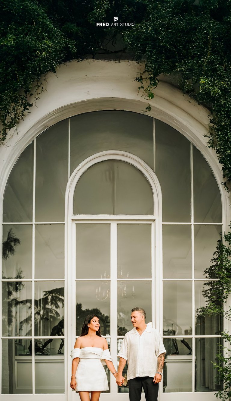 Couple holding hands in front of large white window – engagement photography by Fred Art Studio