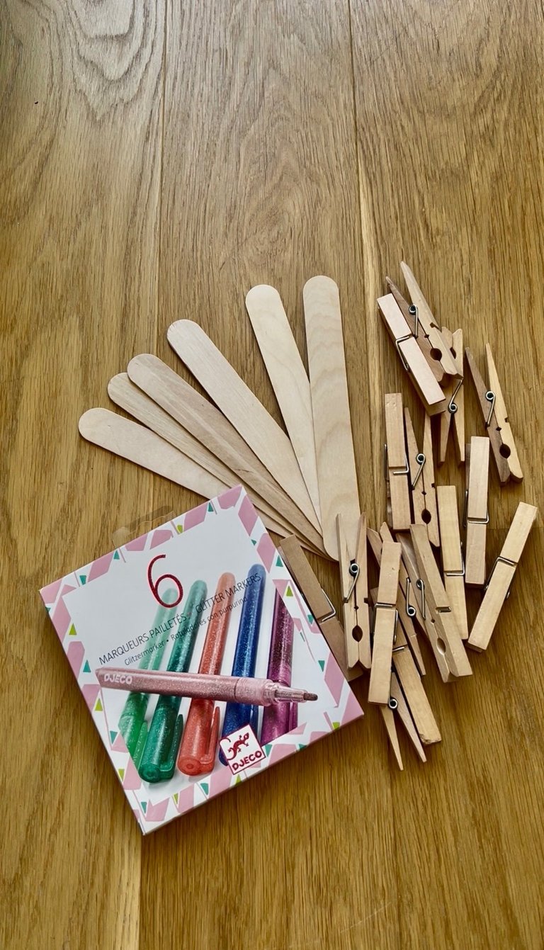 A box of Djeco glitter markers next to wooden craft sticks and clothespins on a table.