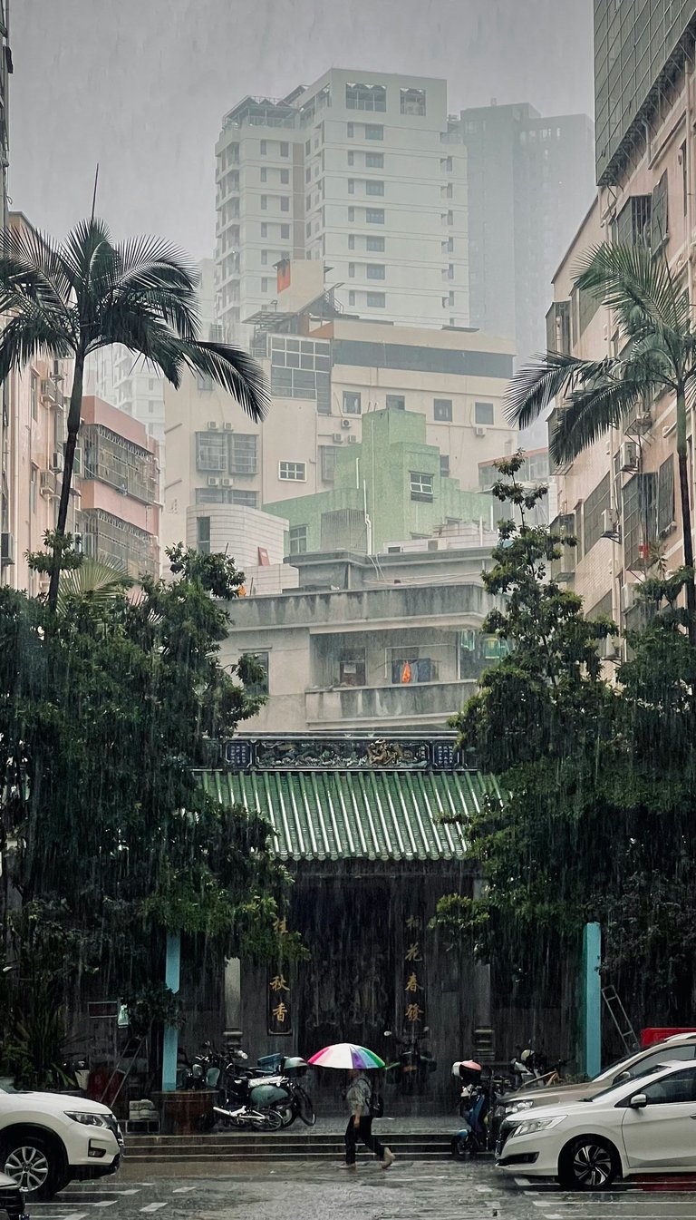 A person with a rainbow umbrella walks past a traditional temple during a heavy rainstorm in a dense city.