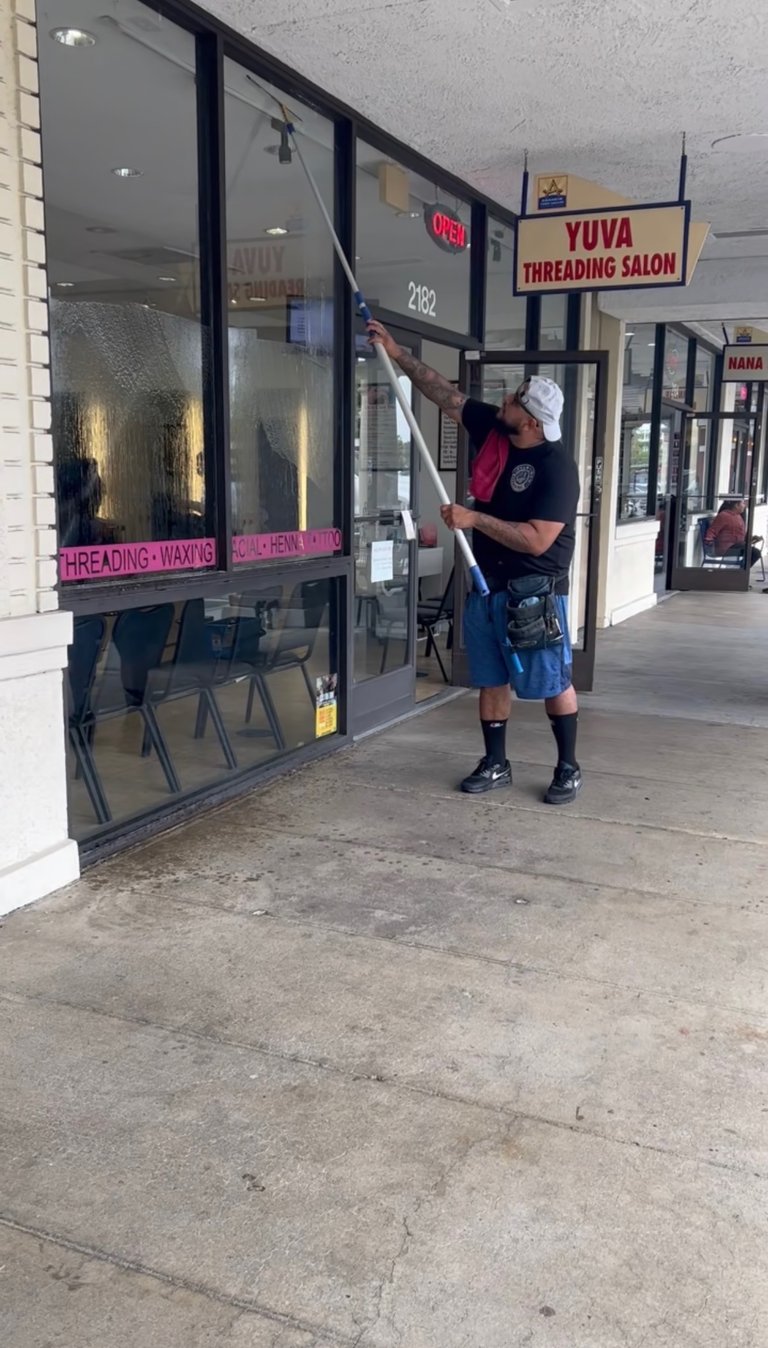 Professional window cleaner using a long extension pole to wash the glass storefront of a local business.