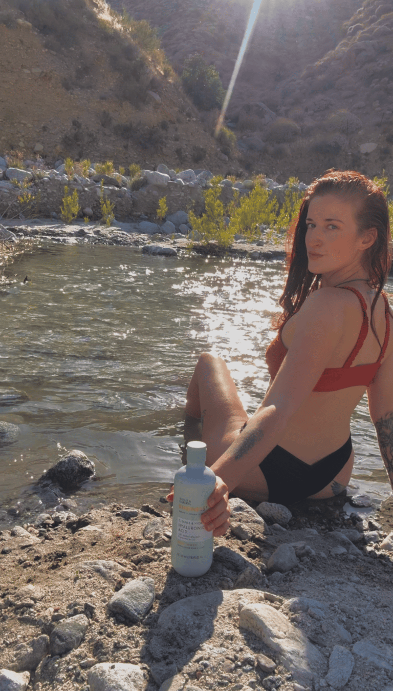 A woman in a red bikini sits by a sunlit river holding a bottle of hyaluronic acid skincare.