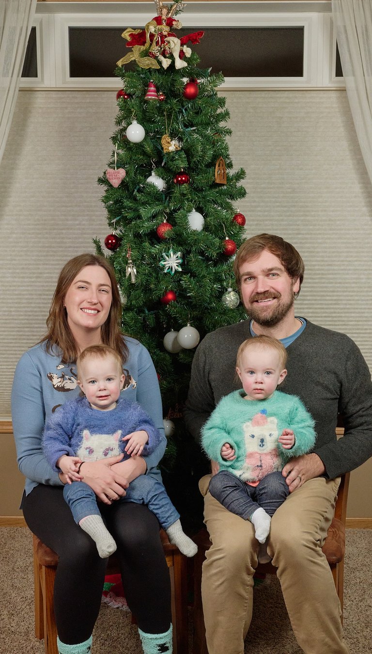 A happy family of four with twin babies sitting in front of a decorated Christmas tree.
