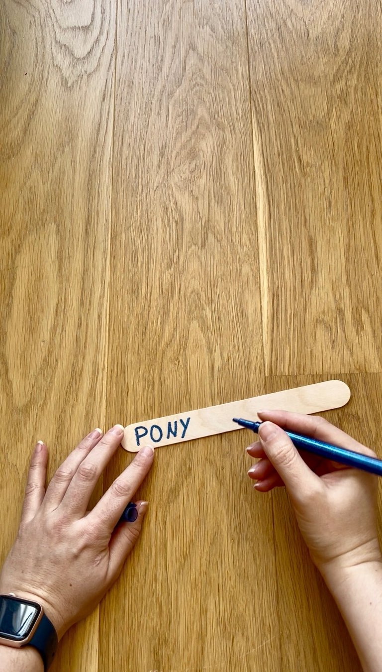 Hand writing the word PONY with a blue marker on a wooden popsicle craft stick.