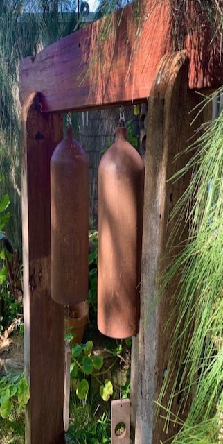 Rustic iron wind chimes hanging from a weathered wooden frame in a lush garden setting.
