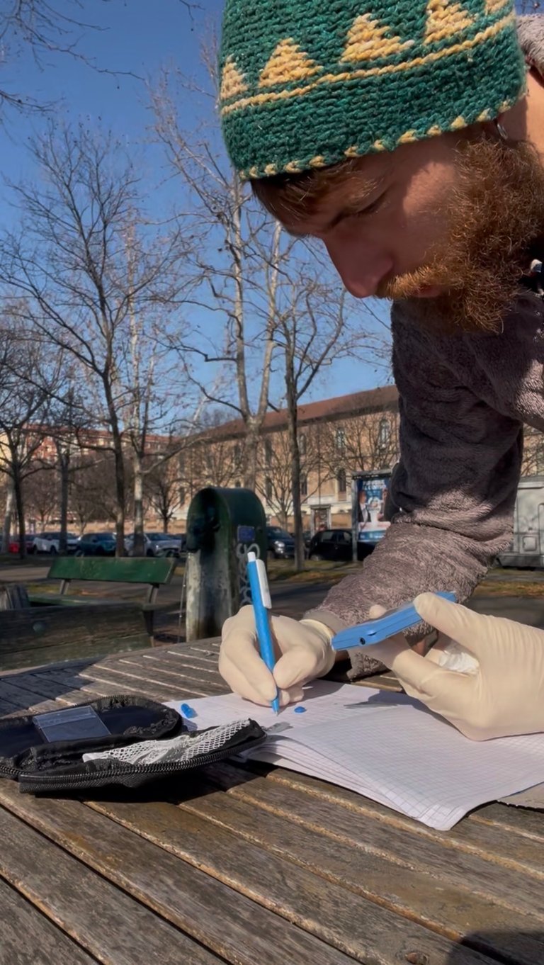 A man with a beard and knit hat wearing medical gloves while writing in a notebook outdoors.