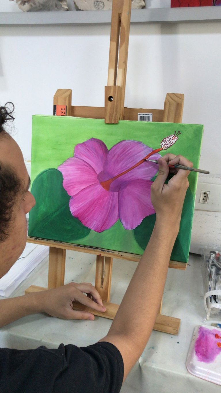 Artist painting a pink hibiscus flower on a green canvas using a fine brush and wooden easel.