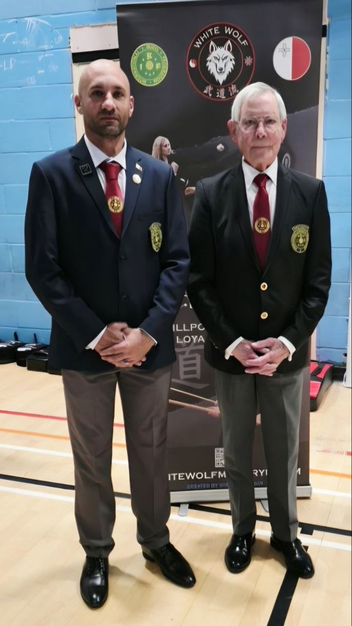 Two karate officials in blazers and ties standing in front of a White Wolf martial arts banner.