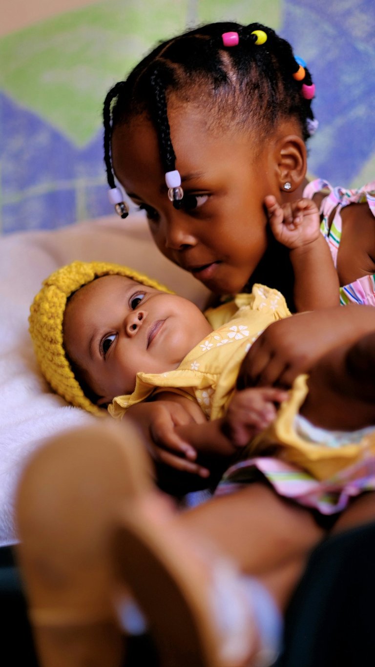 Young Black girl with braided hair gently holding her baby sister wearing a yellow knit hat.