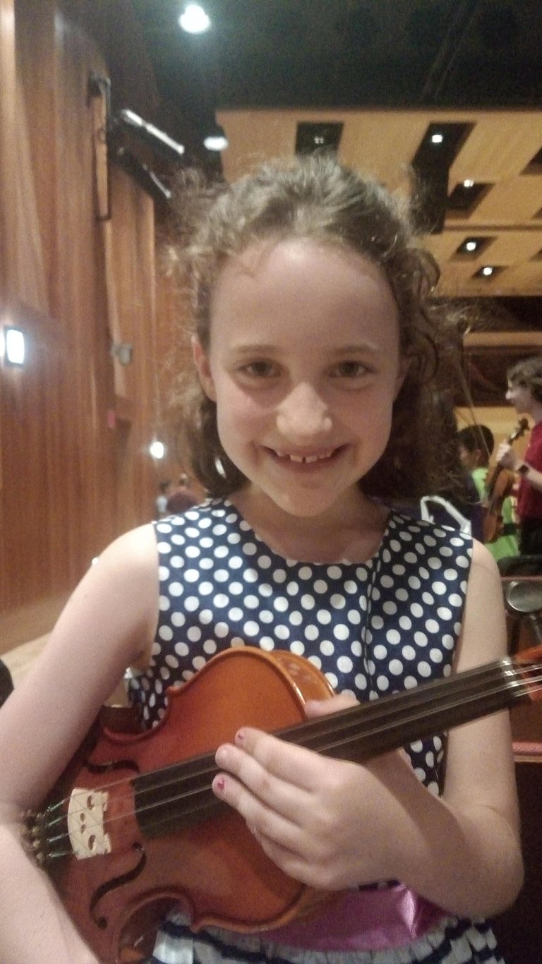 A smiling young girl in a polka dot dress holds a violin during a summer Suzuki Institute.