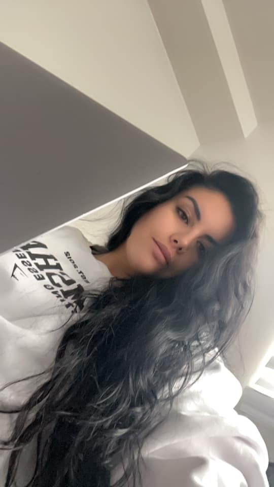 Young woman with long dark wavy hair wearing a white sweatshirt looking into a laptop camera.