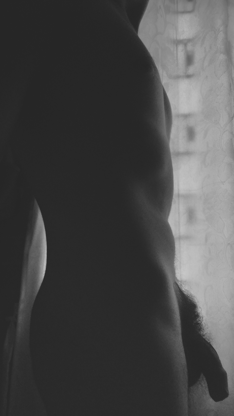 Hombre en blanco y negro completamente desnudo frente a una ventana.