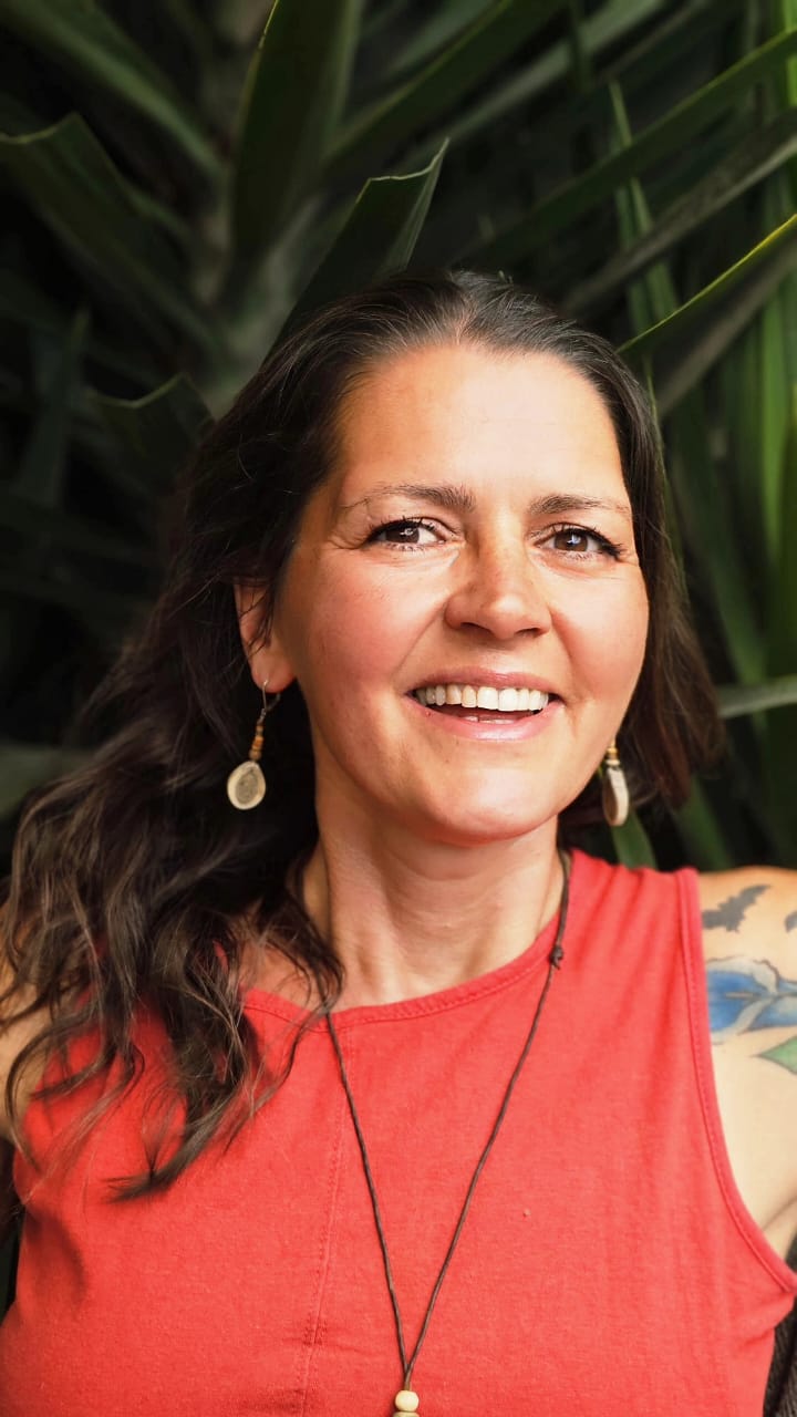 Smiling woman with brown hair and a red top posing against a backdrop of green tropical plants.