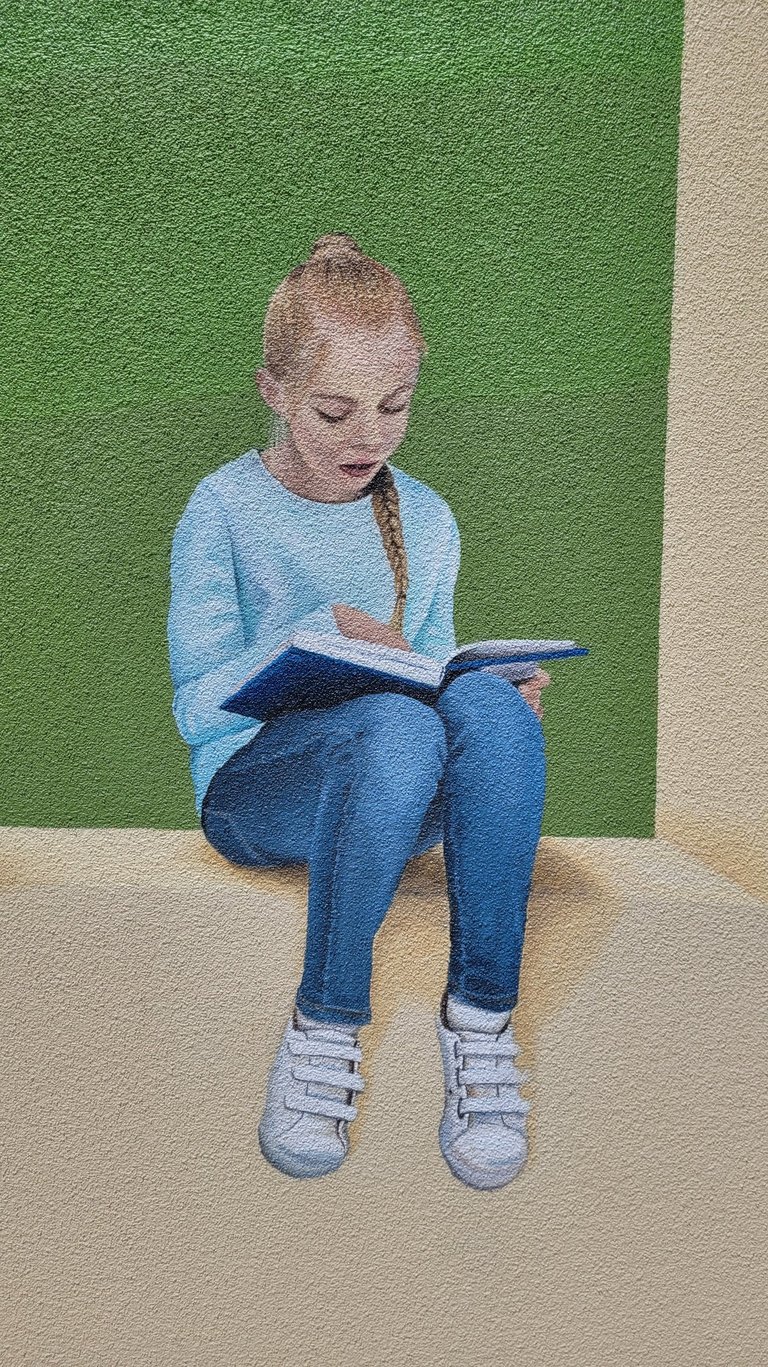 Fresque peinte sur façade. Une fille lit assise sur le bord d'un muret.