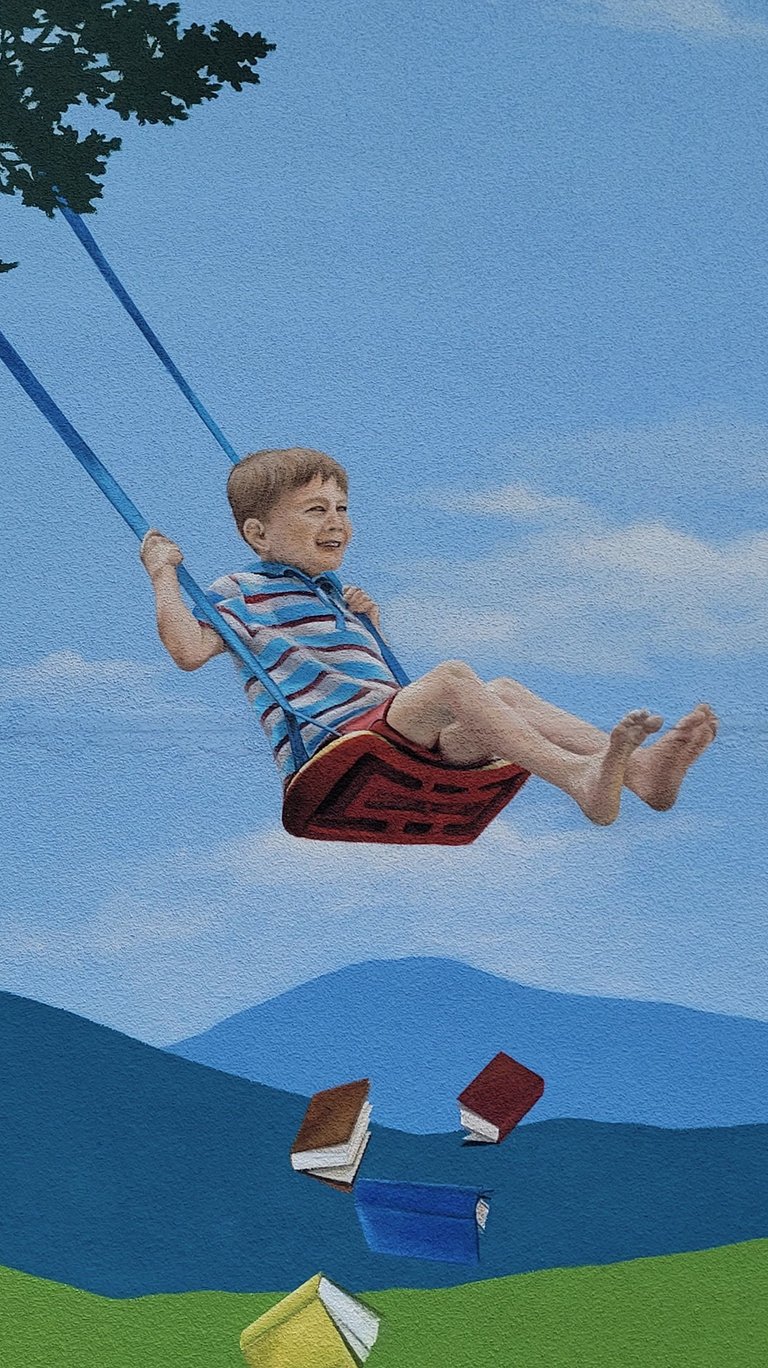 Fresque sur façade. Un garçon joue sur une balançoire au-dessus d'une pile de livres.