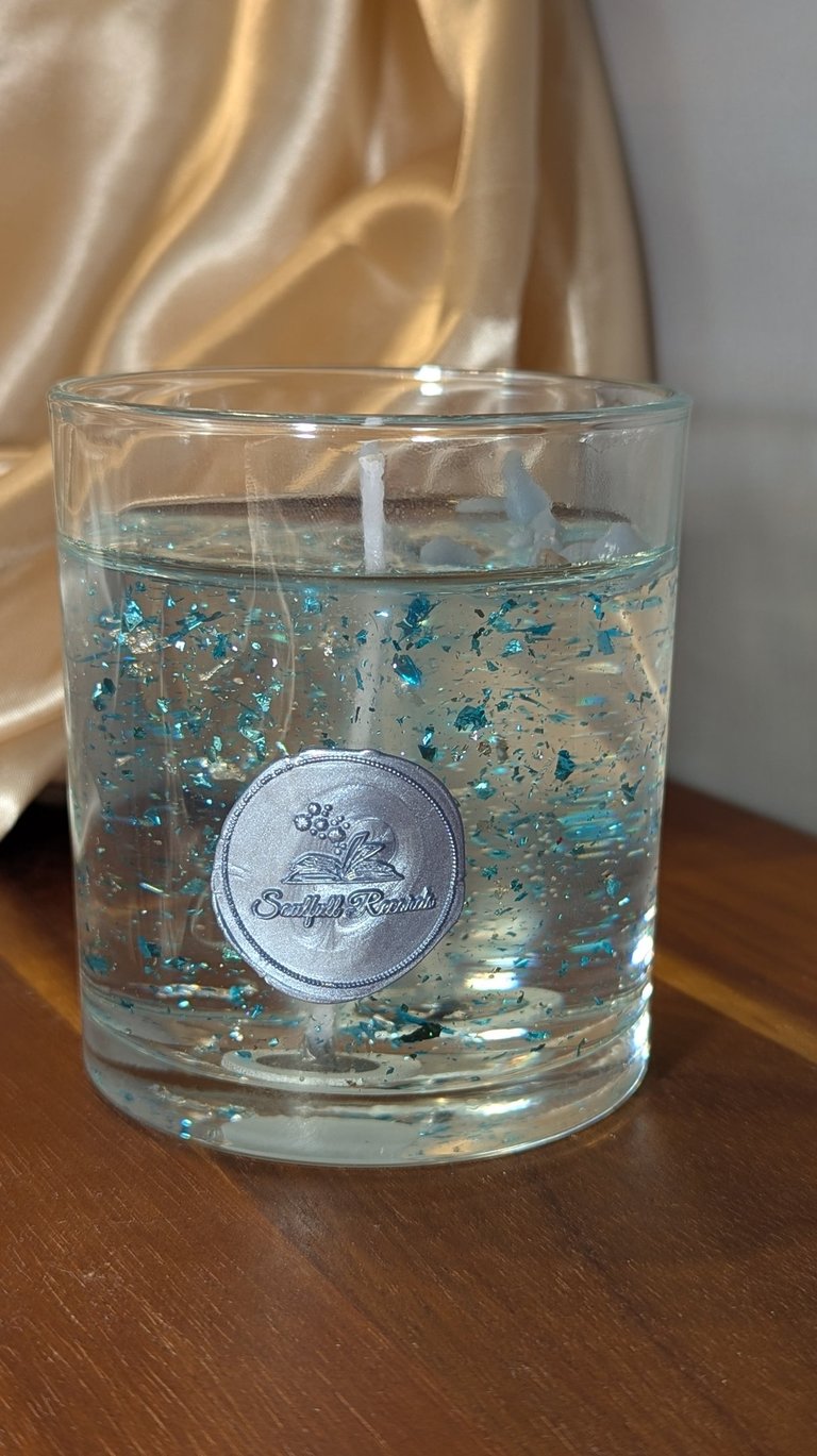 Clear gel candle in a glass jar featuring decorative blue glitter flakes and a silver wax seal logo.