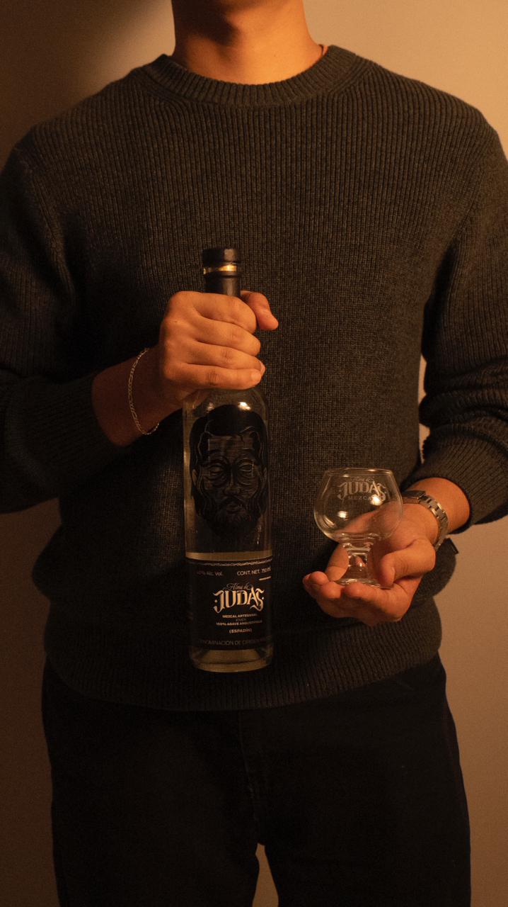 A person holding a bottle of Alma de Judas artisanal mezcal and a small tasting glass.