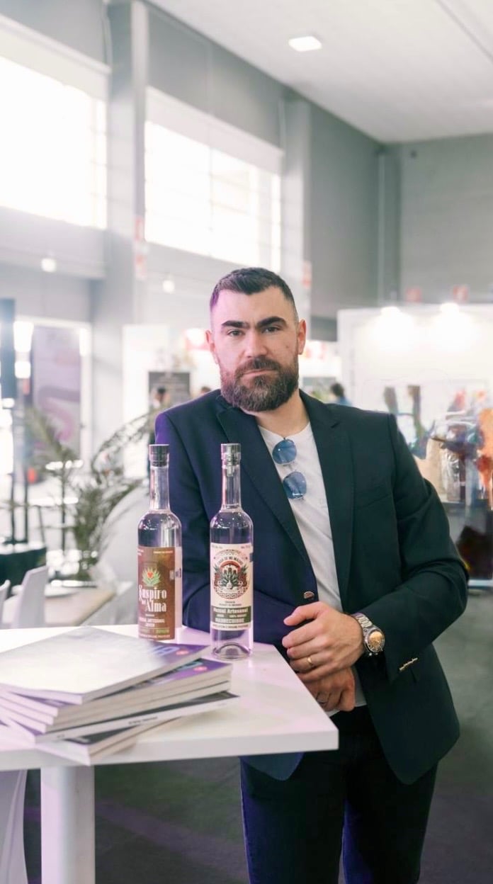 Bearded man in a navy blazer presenting bottles of Suspiro d’Alma artisanal mezcal at a trade show.