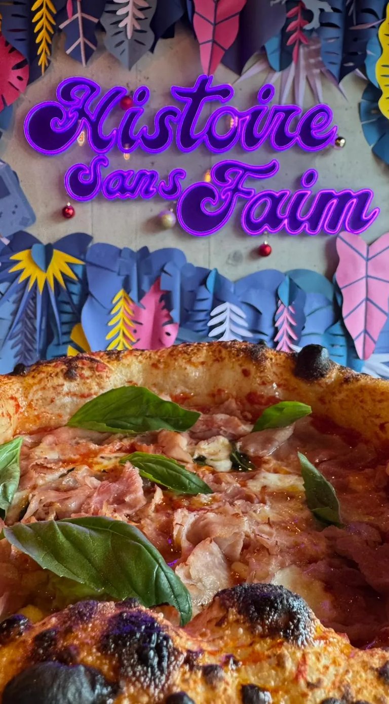 Pizza au jambon et basilic devant l’enseigne lumineuse du restaurant Histoire Sans Faim à Lausanne