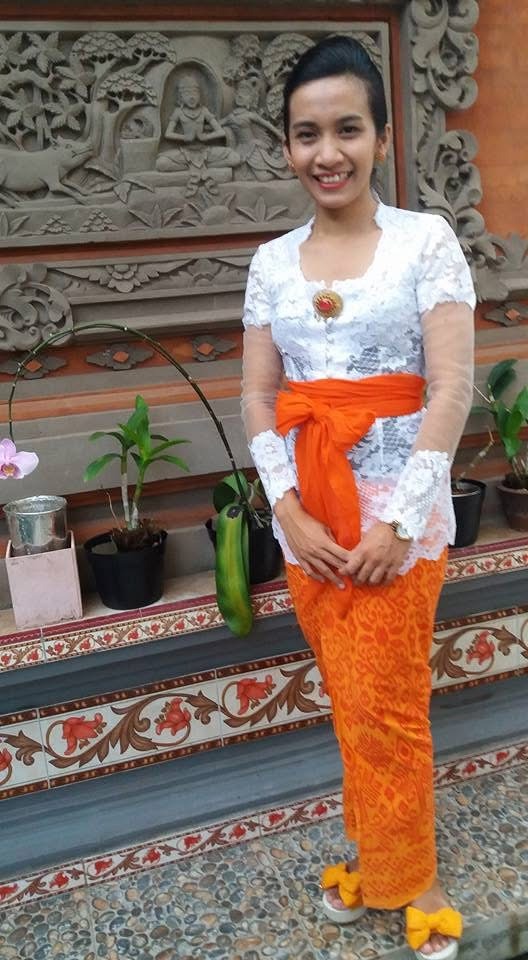 A Balinese woman wearing a white lace kebaya and orange sarong stands before a carved stone wall.