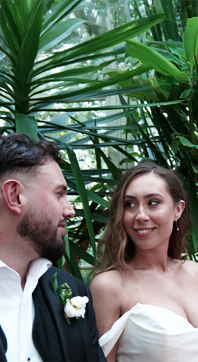 Bride and groom seated together in front of a lush tropical plant, smiling and enjoying their special moment.