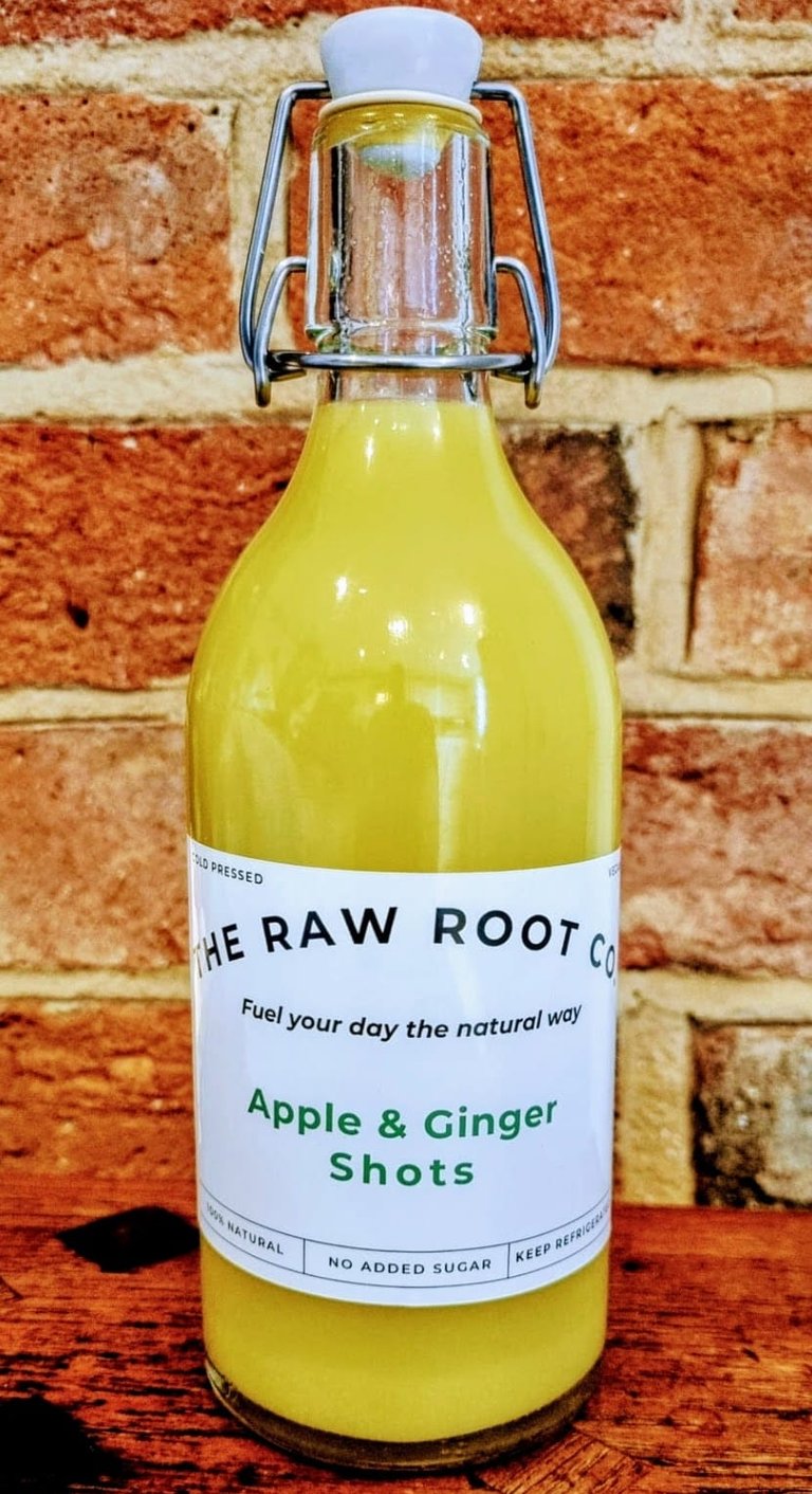 Apple & Ginger health and wellness shots in a glass swing top bottle from The Raw Root Co. Displayed on a wooden table.