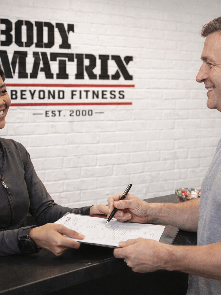 A smiling gym member signs a membership contract at the Body Matrix fitness center reception desk.