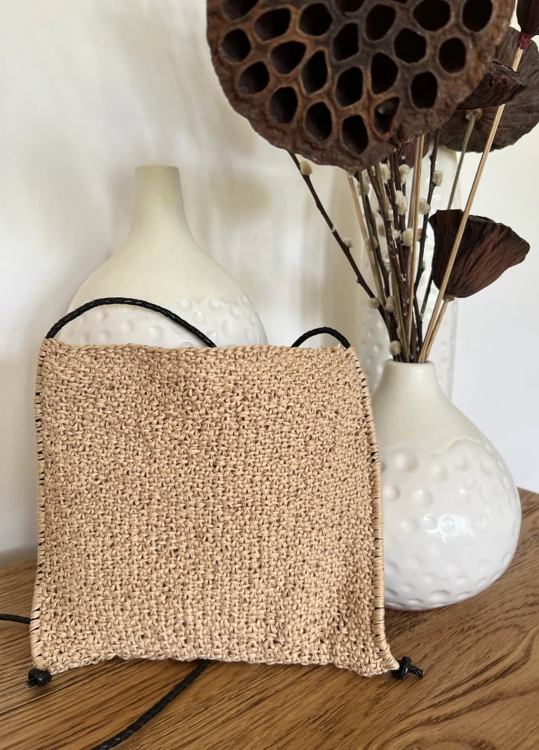 Handknit bag on a wooden table with cream ceramic pots and dried arrangement.