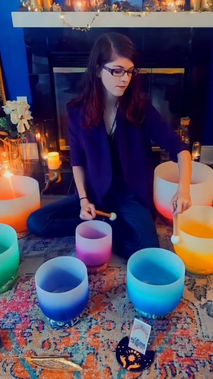 A person seated on a colorful rug is playing crystal singing bowls of different colors and candles 