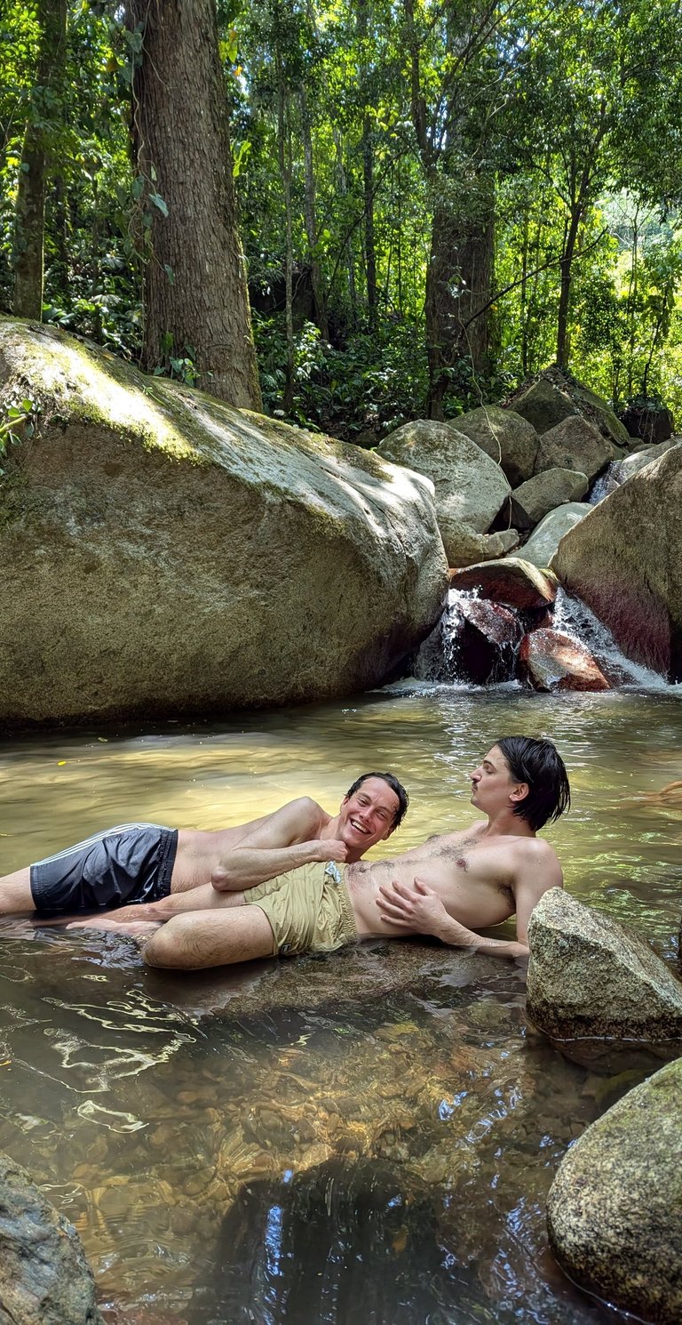Friends relaxing in a natural jungle waterfall pool surrounded by tropical trees and large mossy rocks.