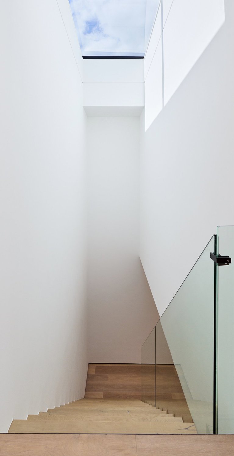Modern des staircase with glass railing leading to a bright skylight in a minimalist white interior.