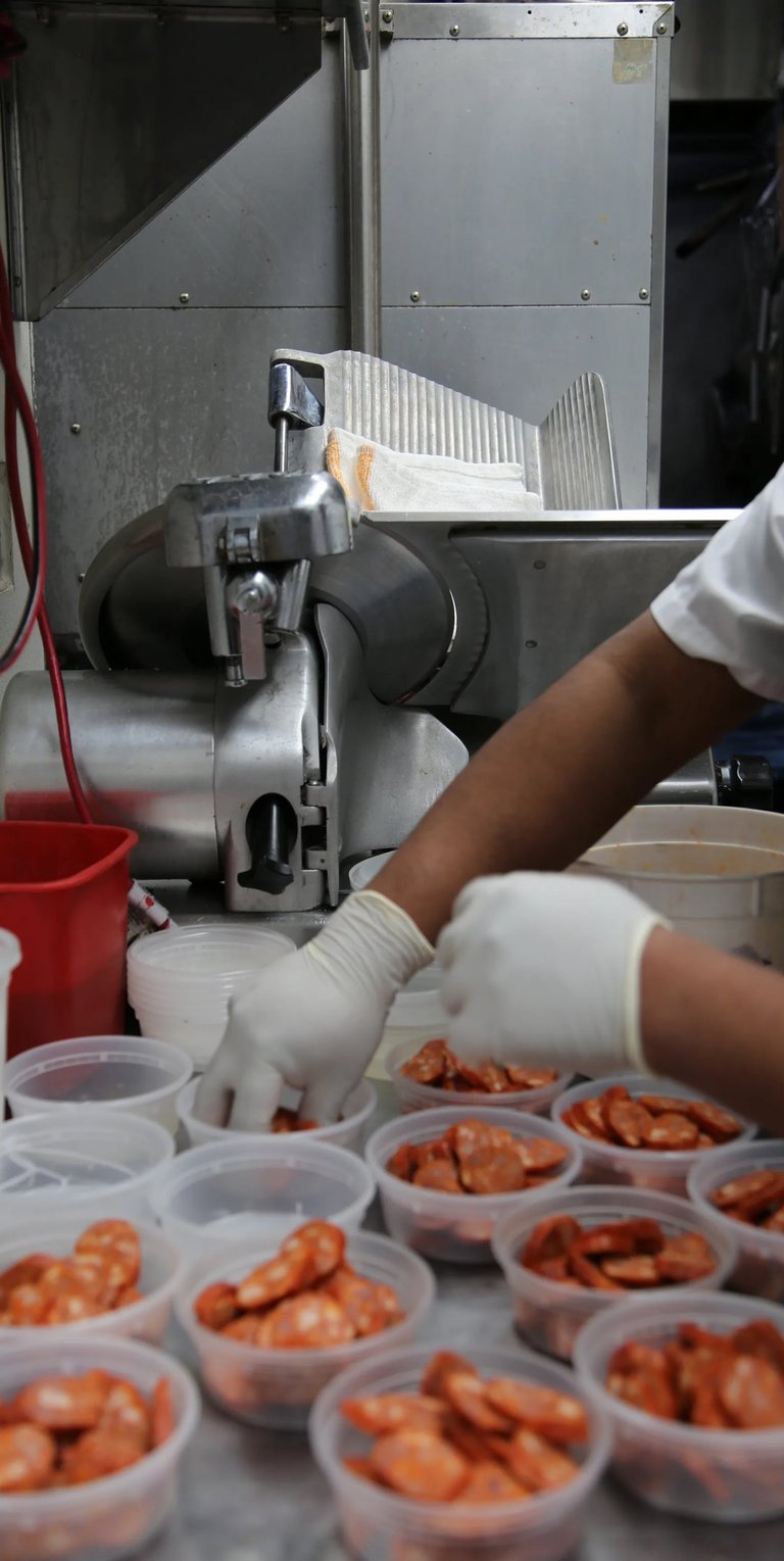 Gloved hands placing sliced chorizo into containers in a professional kitchen.