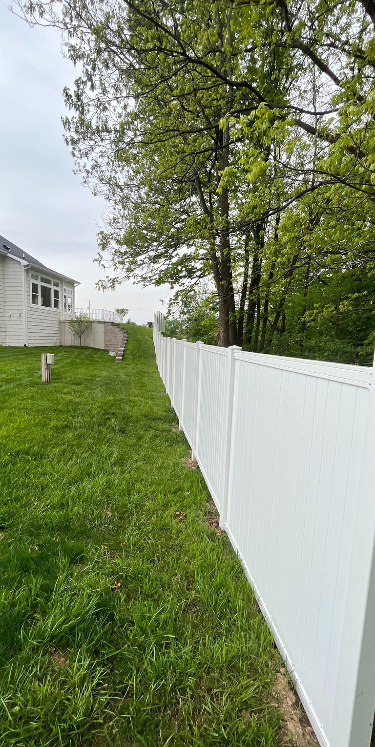 White vinyl privacy fence installed in a green backyard bordering a wooded area next to a white house.