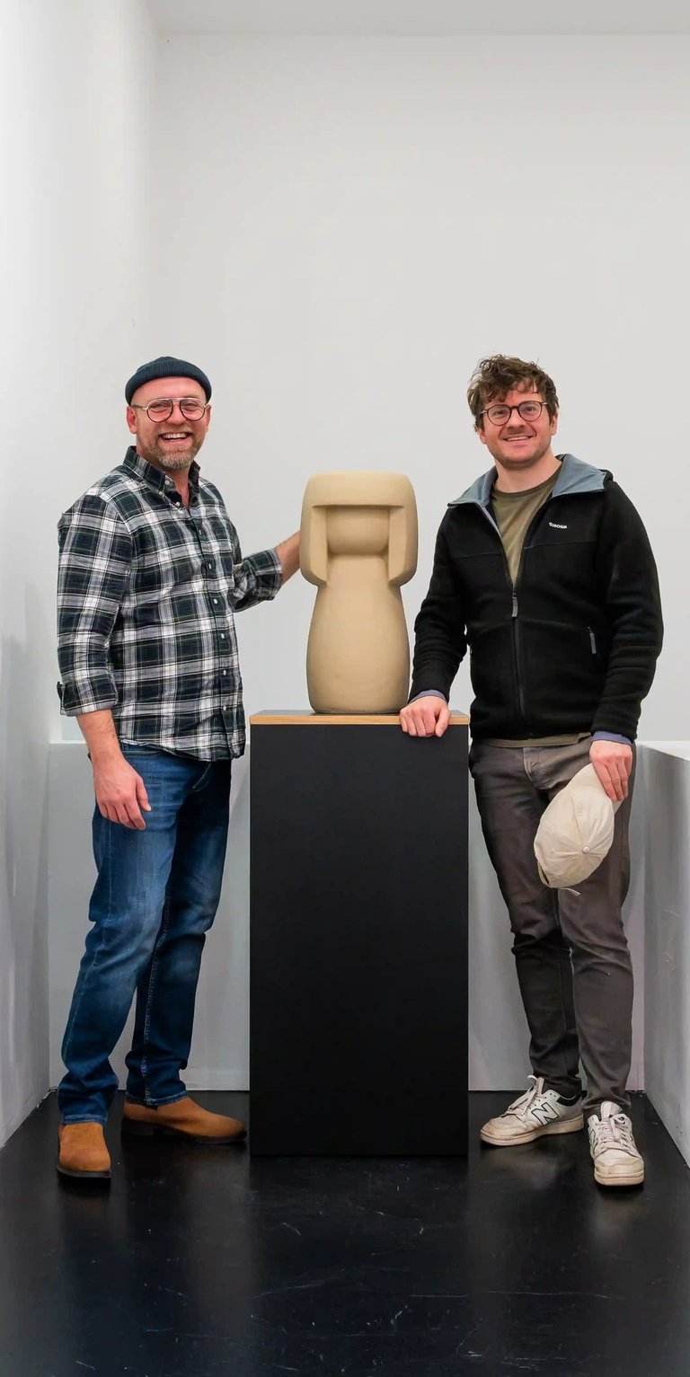 Richard und Felix stehen neben einer Kokeshi, die auf einem Podest steht