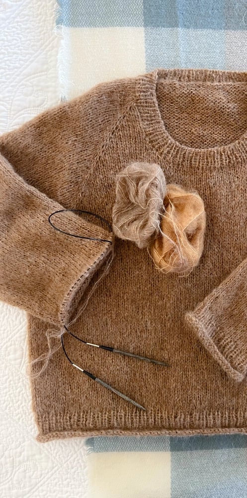 Unfinished beige hand knit sweater lying on a checked blanket with knitting needles and yarn.