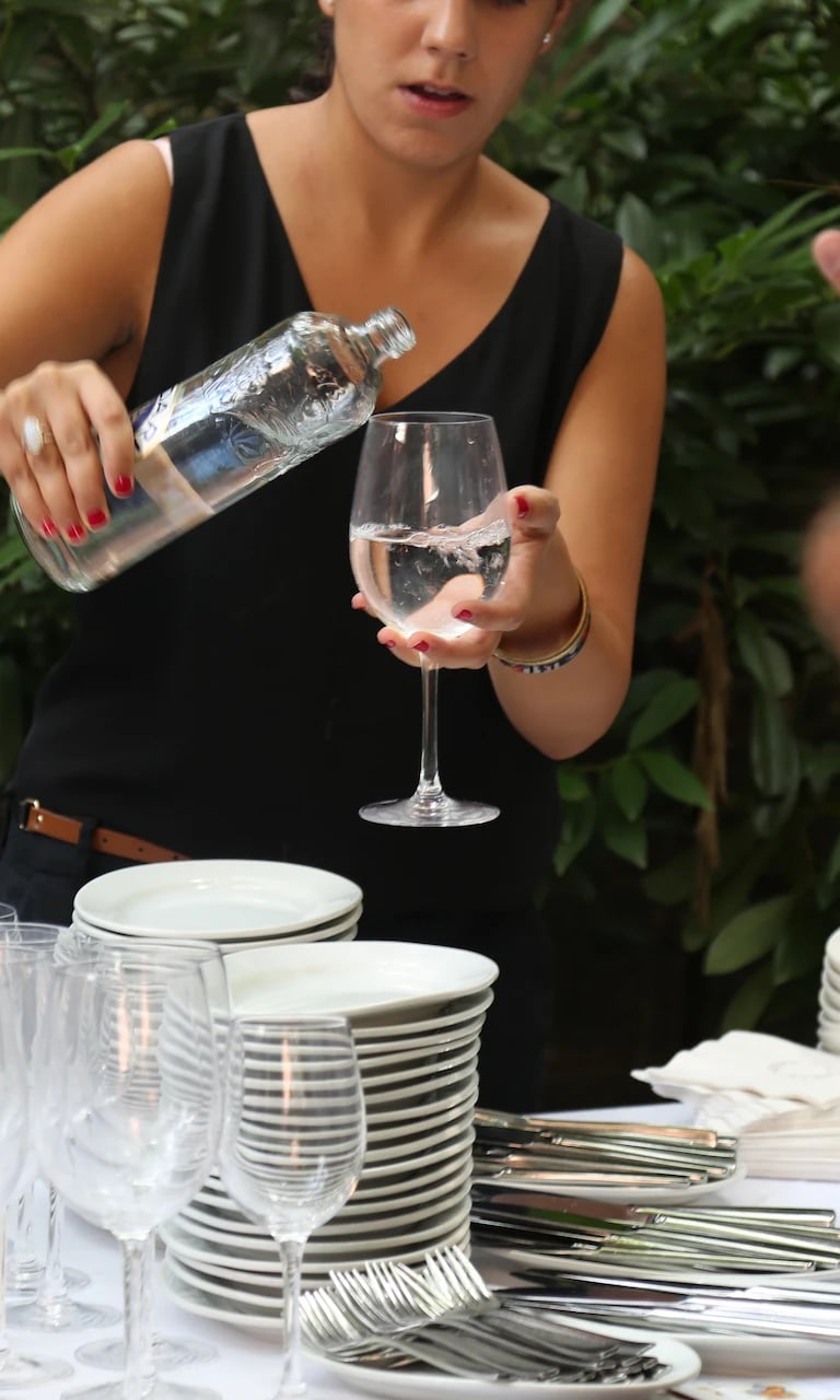 Woman dressed in black pouring water into a glass next to a table set with plates and glasses in a restaurant.