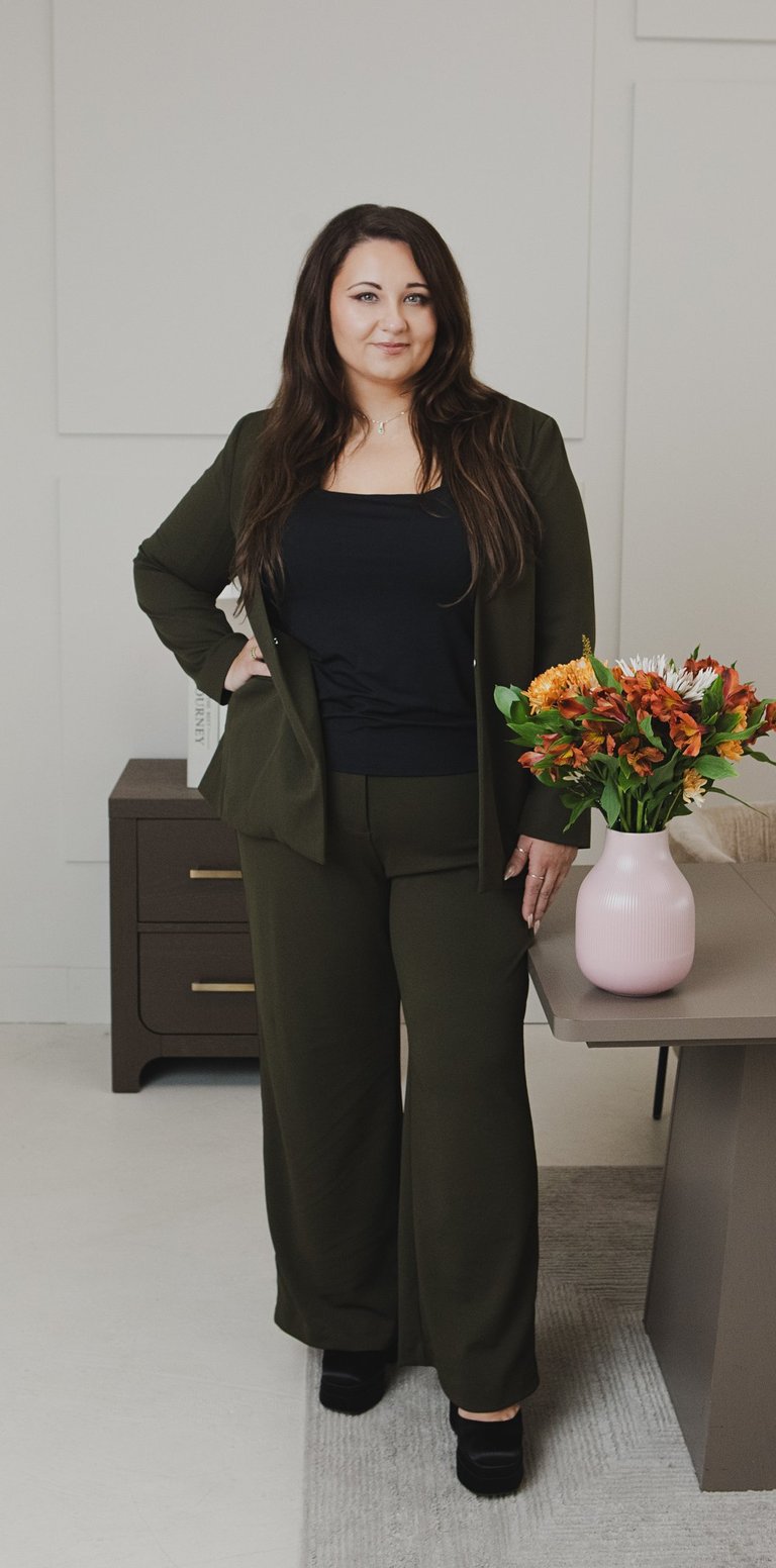 Adriana in dark green blazer and trousers standing by a desk with autumn flowers.